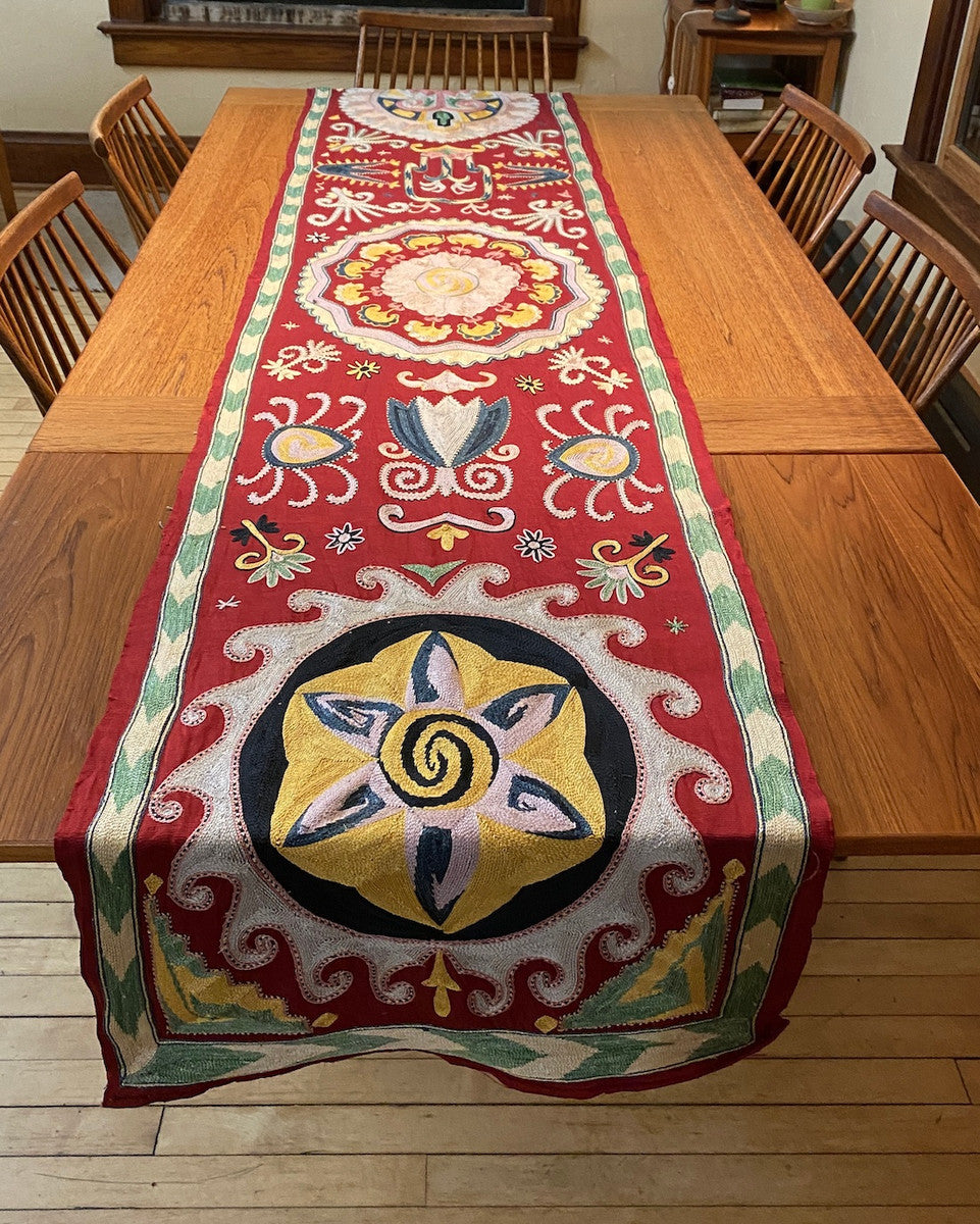 Hand Embroidered Silk Table Wall Hanging Uzbekistan Embroidery colors: chalky leaf green, pinkish cream, pale bluish cream, chalky pale pink, pale violet cream, pale yellow, Prussian blue, gold, and black that sit atop a silk ground cloth of deep red.